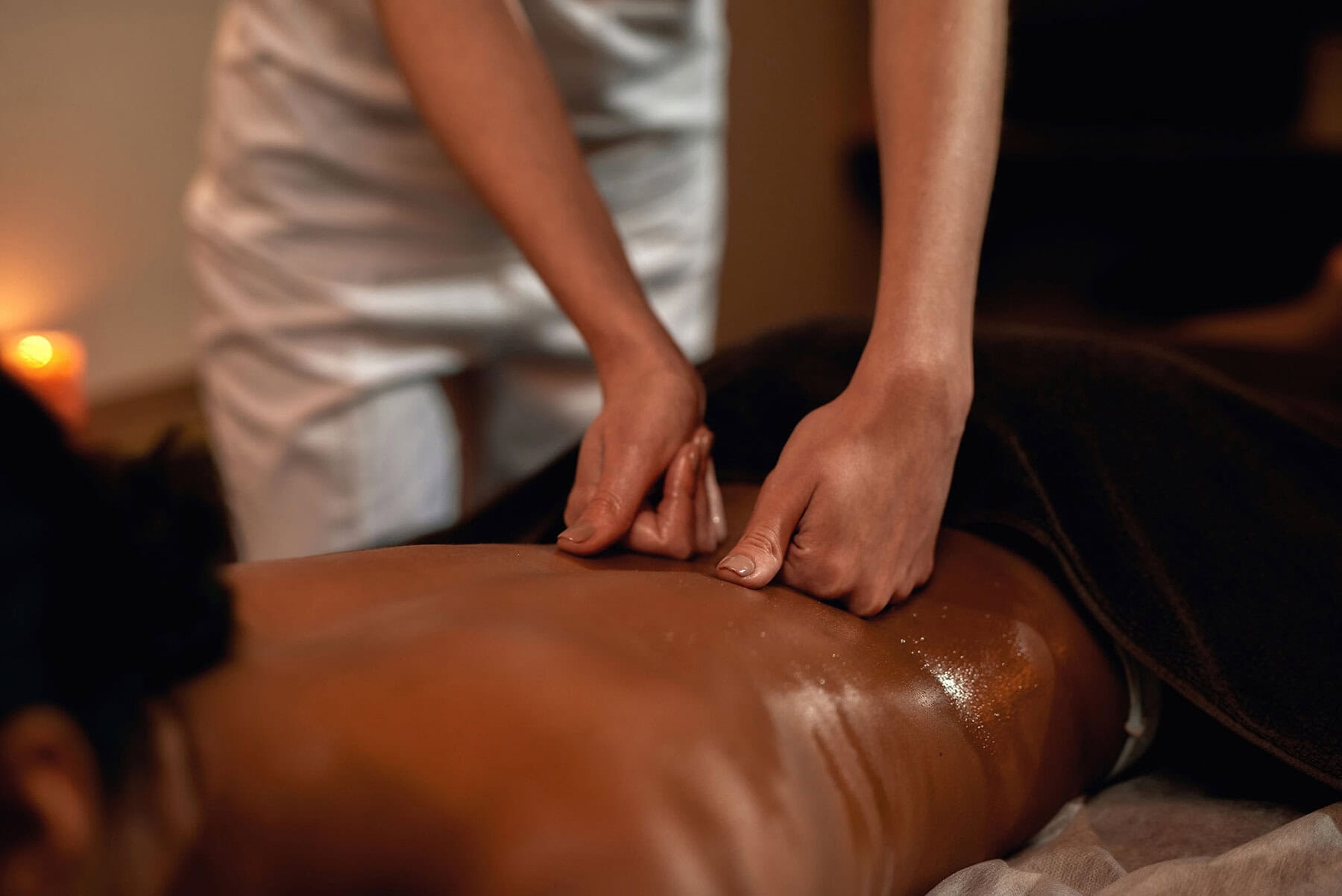 Partial image of female masseur doing back massage of relaxing woman in spa salon. Young black girl client lying naked under towel on massage table. Concept of rest with spa treatment